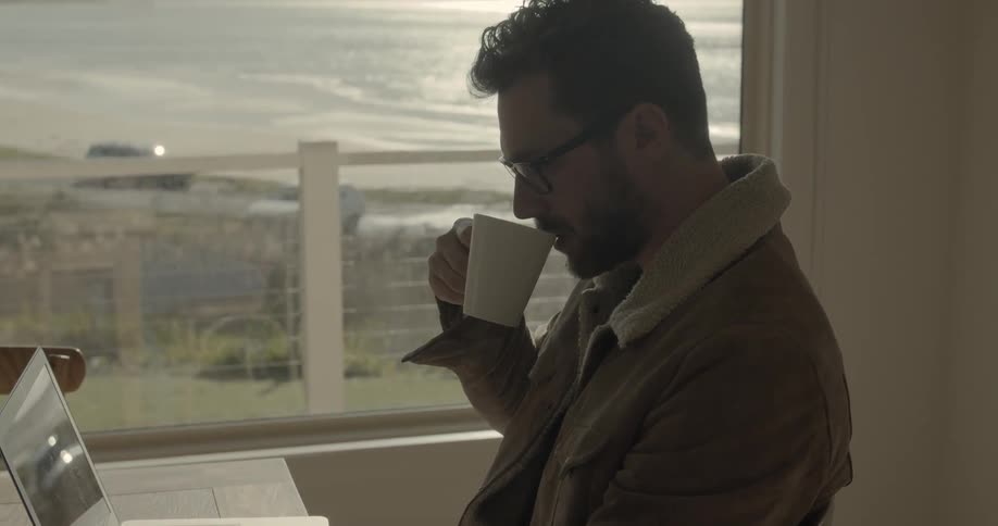 Man sits at the table and uses laptop while drinking coffee indoors during daylight