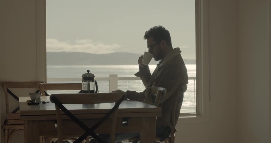 A man sitting at a table in front of a large window drinking coffee from a mug