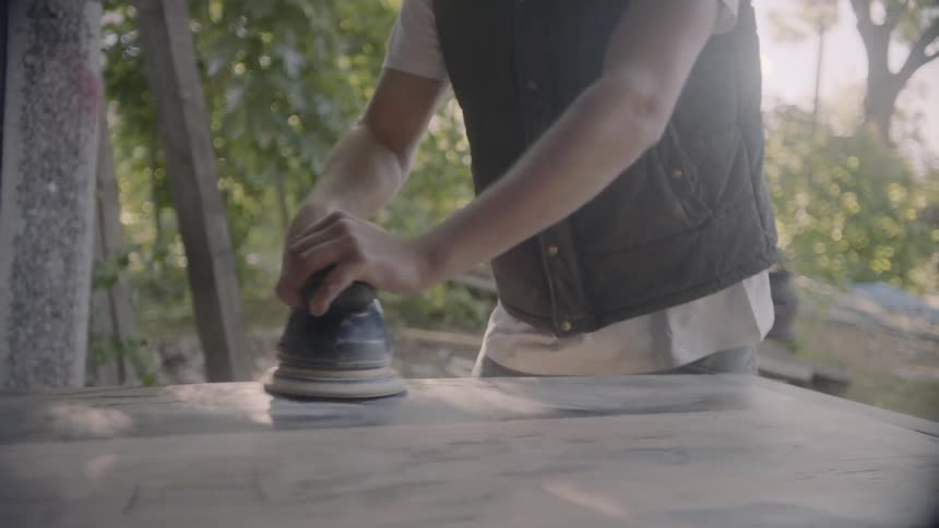 Blonde male sanding a slab of wood outdoors near trees