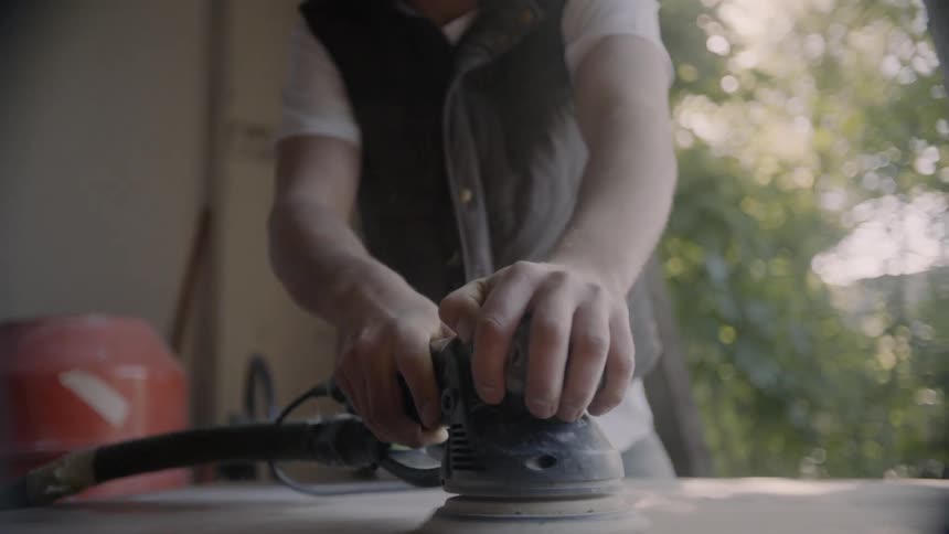 A man is holding a rotary tool that he is using to polish the surface of a piece of wood that hes working on