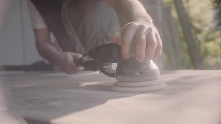 A man is holding an electric rotary hand tool that he is using to polish the surface of a wooden board that is located in a garage work space with plenty of natural lighting