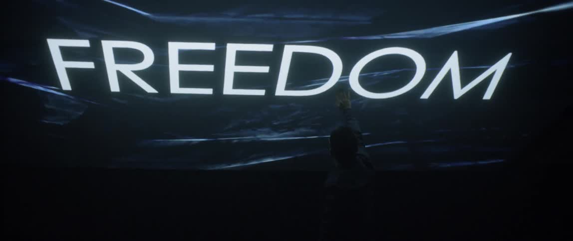 In a dark room the word freedom is displayed on a large screen while a man in glasses waves his hand back and forth in front of it