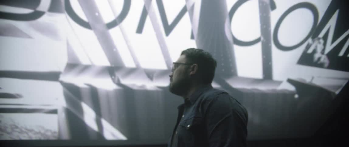 A bespectacled man in a denim shirt stands in front of a screen displaying the lords prayer in more than one language in stylized form which blanks out before returning