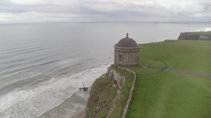 Waves break and splash as they come to shore on the beach below the domed stone round temple on a cliff