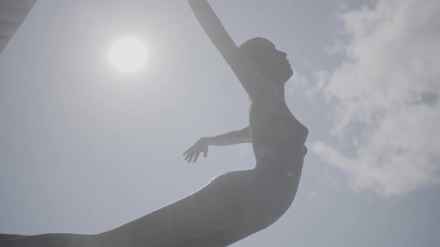 A statue of a woman with her hands outstretched to her sides is seen below a blue sky with the sun shining bright and a few clouds