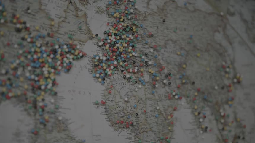 A vintage style map of the world is posted on a wall and has hundreds of colorful pins stuck into various parts of the map all over the world