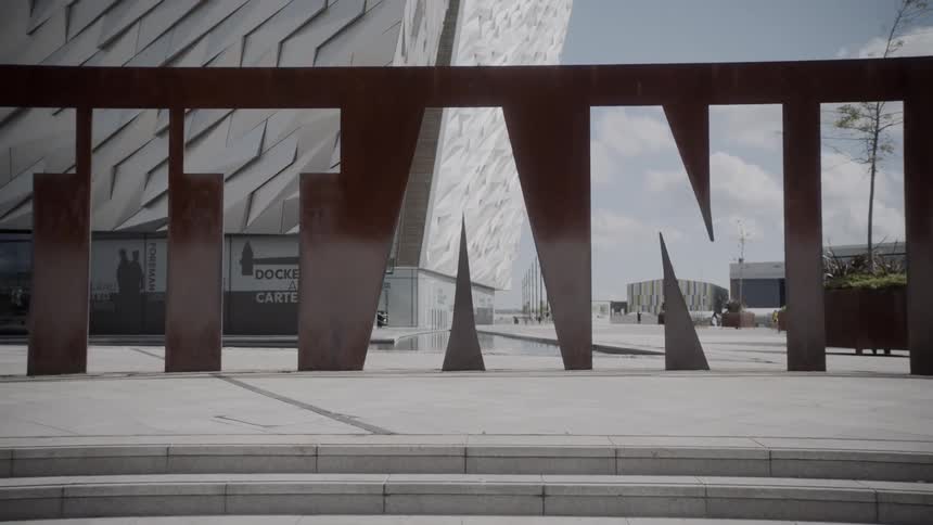 A thin wall is standing as a mural in front of an elaborate building and the mural has letters carved out of it to form open spaces that spell the word titanic