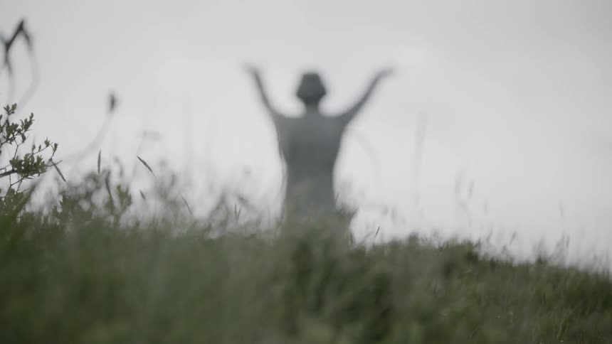 Grass blowing in the wind with a statue of a person with their hands up in the background
