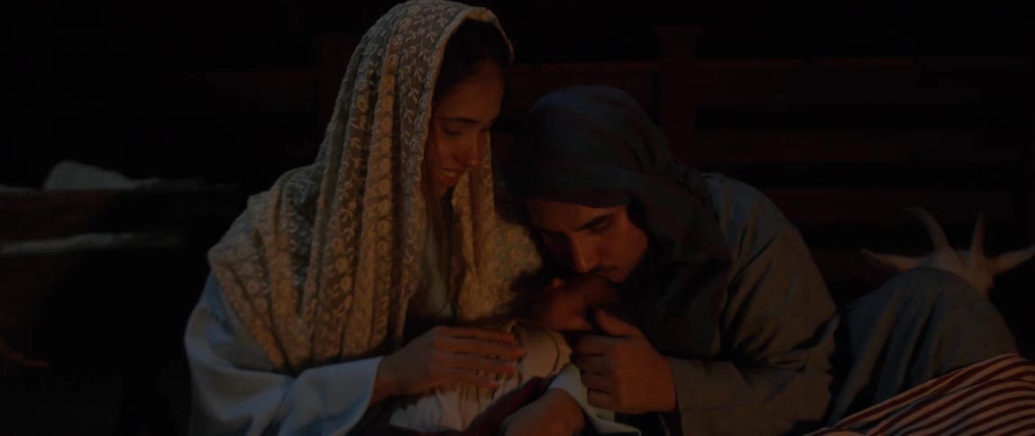Woman holding baby sits with man touching and kissing the baby next to the fire at night