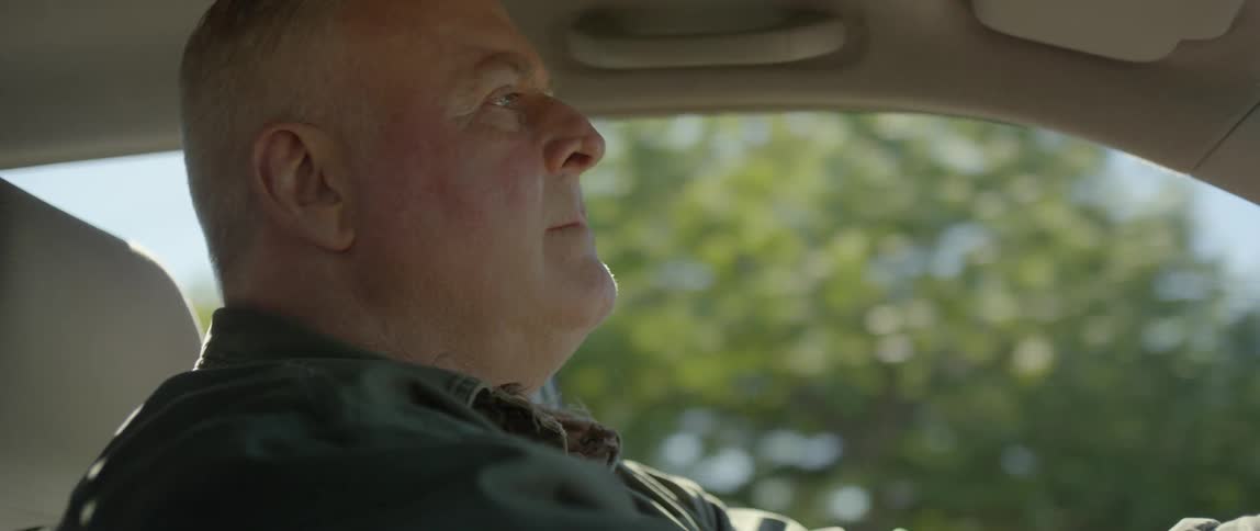 A white older male is driving a car down the road when something catches his attention causing him to gaze over to it