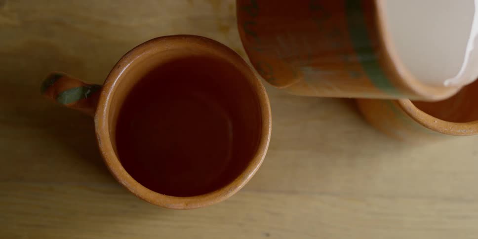 A pale liquid spilling onto the table while being poured into an orange mug