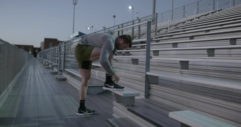 A white man ties his running shoe while standing on bleachers