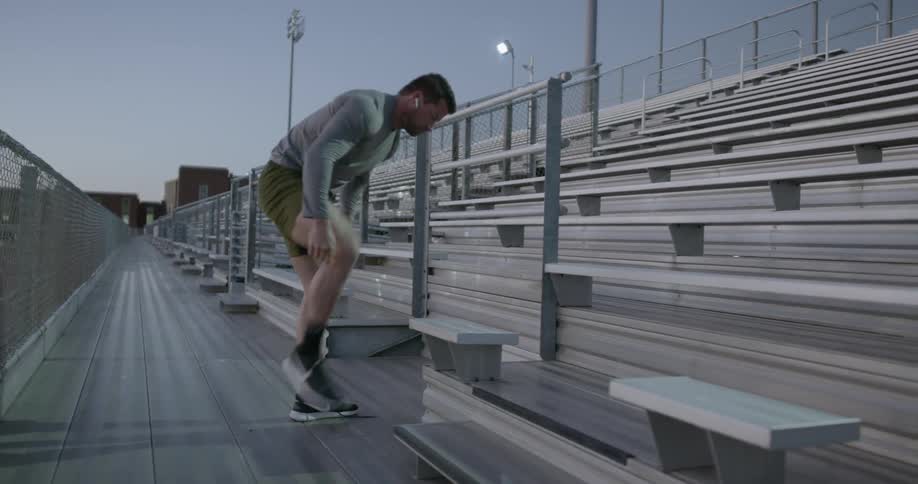 A man wearing earbuds stepping on bleachers to tie his shoes