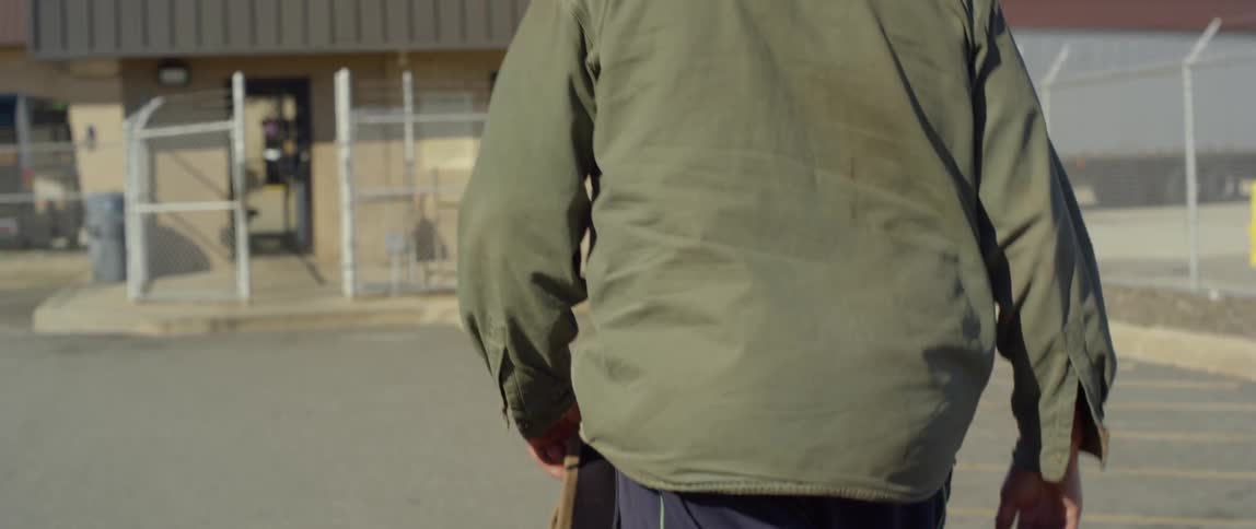A man carrying a beanie hat walks toward a motorcycle parked next to a fence at a rest stop