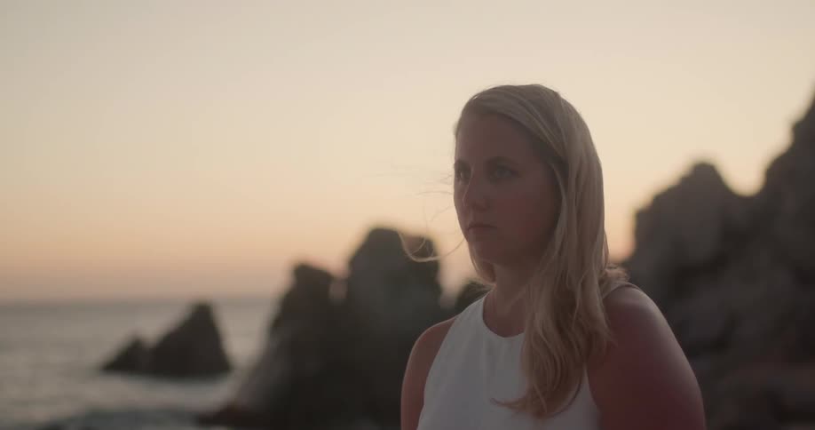 A blonde woman stands on the shore near ocean waves splashing up on the rocks at sunset