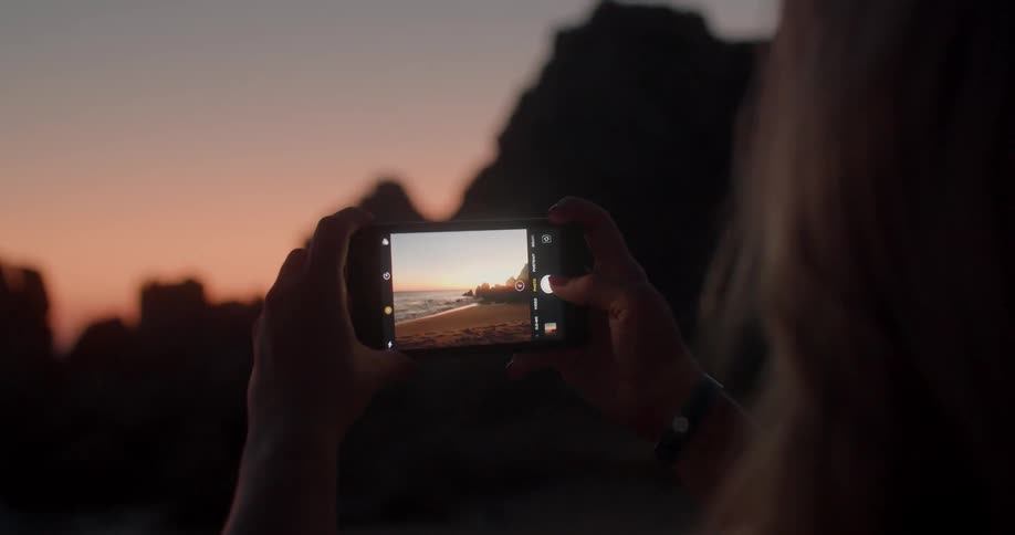 A woman holds up a smartphone and takes a picture of a beach