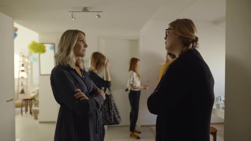 Two women having a discussion in a house while other women talk nearby