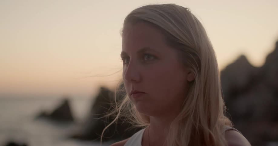 A young white female appears to be deep in thought with water and jagged rocks behind her
