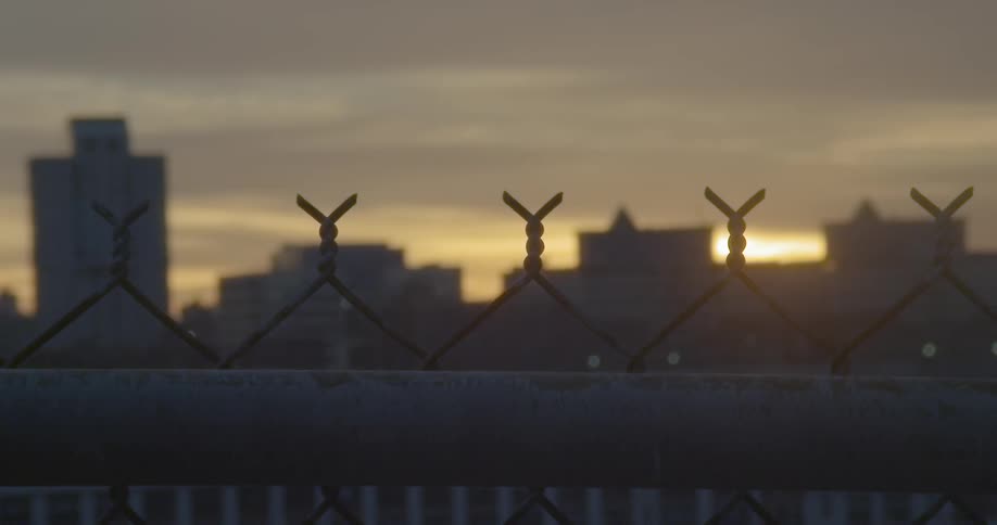 Top of chain link fence with sun rising behind buildings