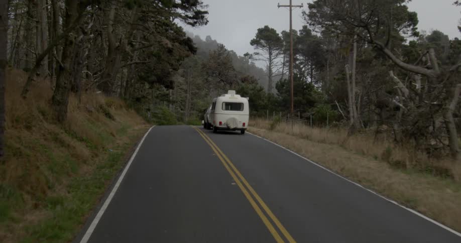 A pickup truck towing a trailer travels on a rural tree lined road