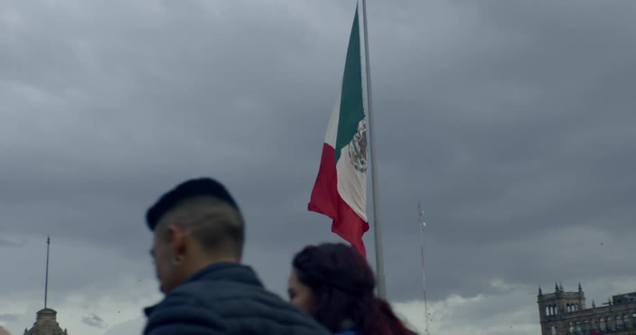 A mexican flag on a flagpole moves in the breeze as people walk by