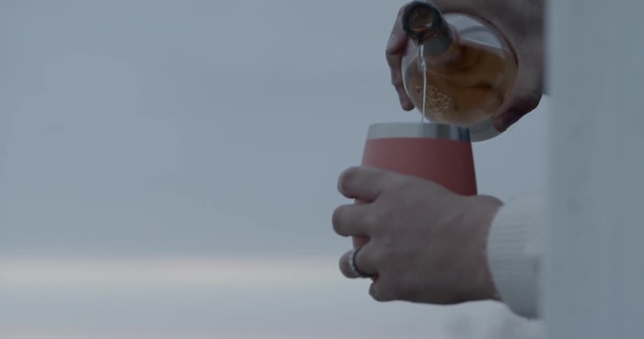 A man holds a glass bottle and pours a drink into a cup hes holding