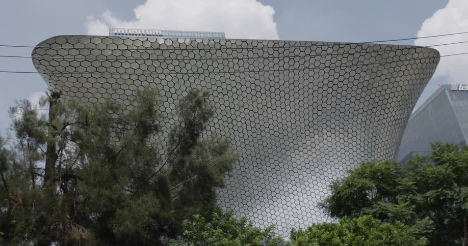 A structure covered in hexagons near a large building and trees