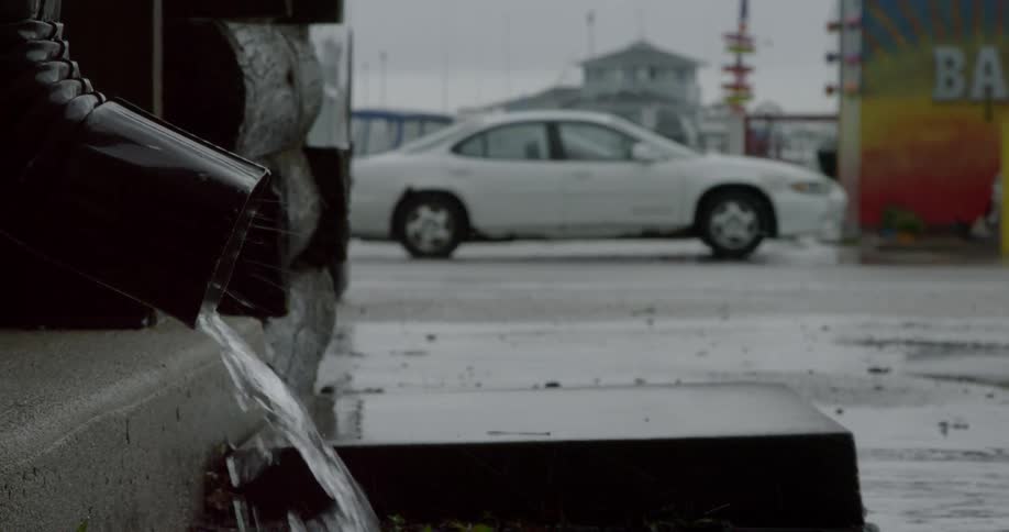 Water pours from a downspout attached to a building with a white car parked in a parking lot behind it