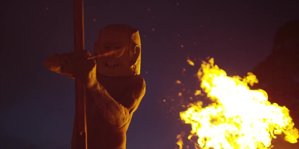 Man in a mask aims an arrow in front of a bonfire