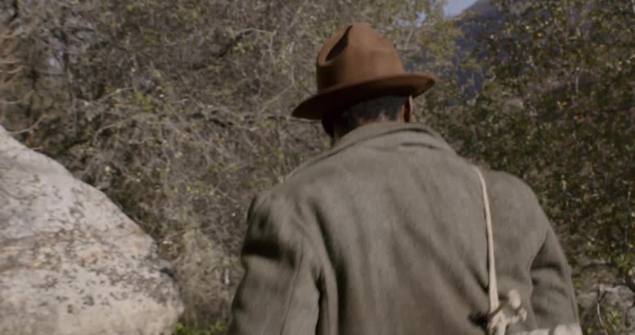A man in a hat carries a duffel bag filled with camping equipment and walks through a tree lined path in a mountainous area