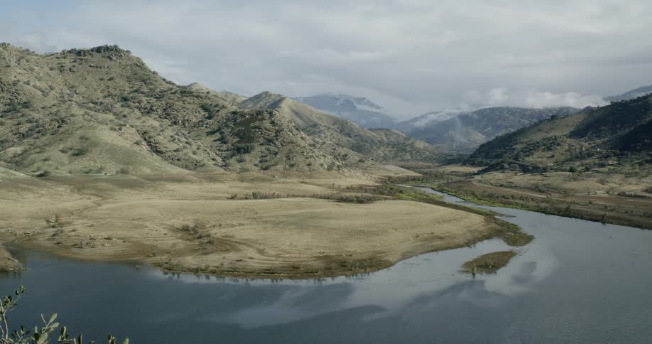A still lake at the bottom of a valley surrounded by mountains sparsely littered with bushes under a cloudy sky