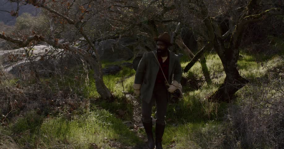A man wearing outdoor gear and carrying camping equipment walks on a path
