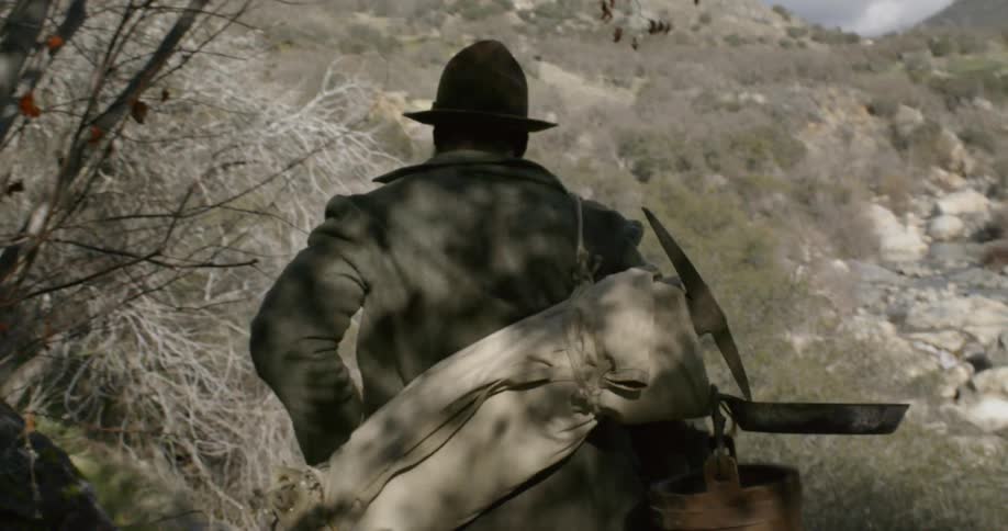 A man in a hat carries a duffel bag filled with mining and camping equipment and walks through a tree lined path in a mountainous area