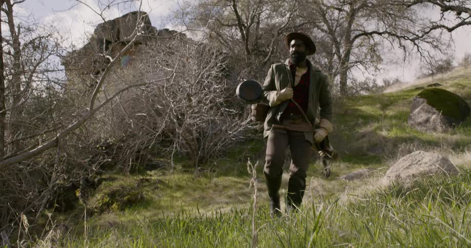 A black male hiking through a grassy area as he carries items