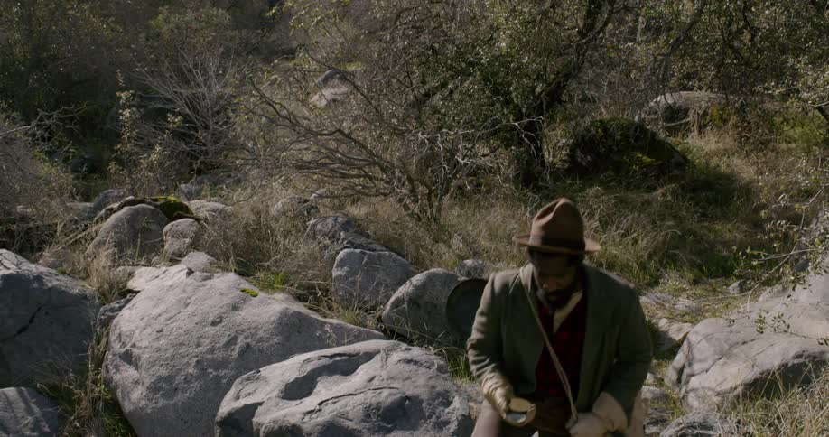 A man in a hat carries a duffel bag filled with mining and camping equipment and walks through a tree lined path in a mountainous area while looking at a compass