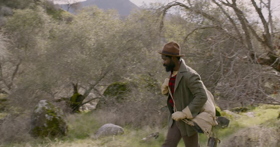 A black man wearing a brown hat and carrying a rucksack walks through the woods