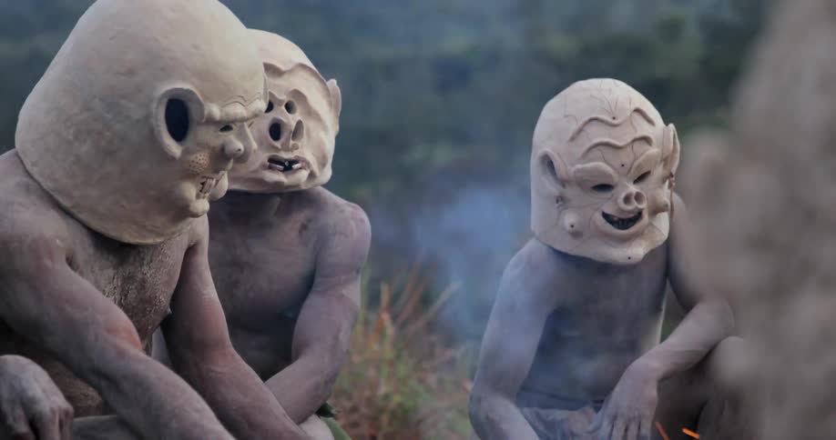 Tribal men in masks gather around an outdoor fire
