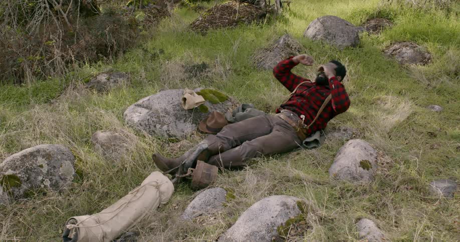 A man sitting in a grassy field looks very tired and then lies down