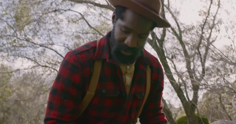 An older black man in suspenders is sorting through objects on a blanket on the grass with trees behind him