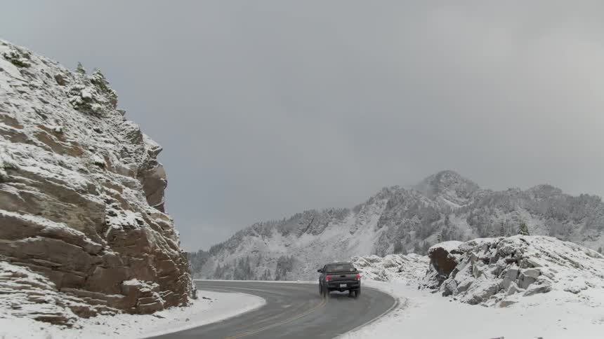A pickup truck driving on a winding road with mountains and snow in the background