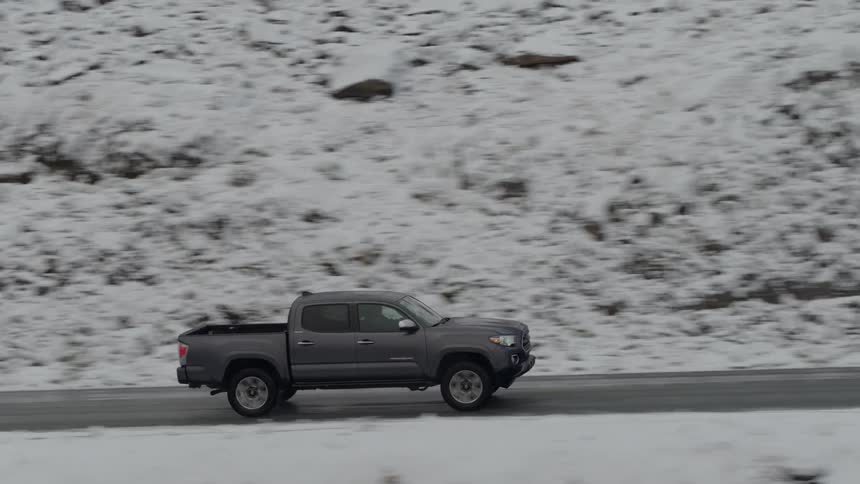A pickup truck with headlights on travels on a mountain road with snow dusted slopes