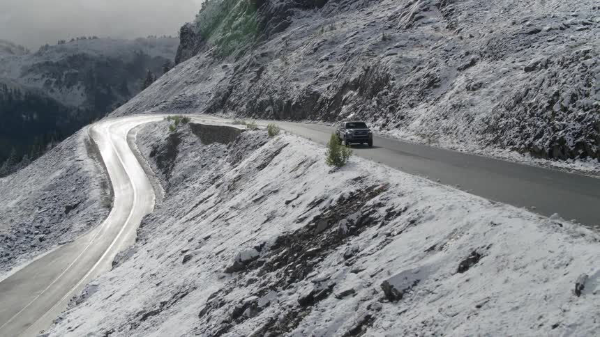 A pickup truck travels on a mountain road with slopes dusted with snow