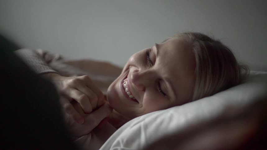 A woman laying in bed smiling and kissing the hand of a man lying next to her