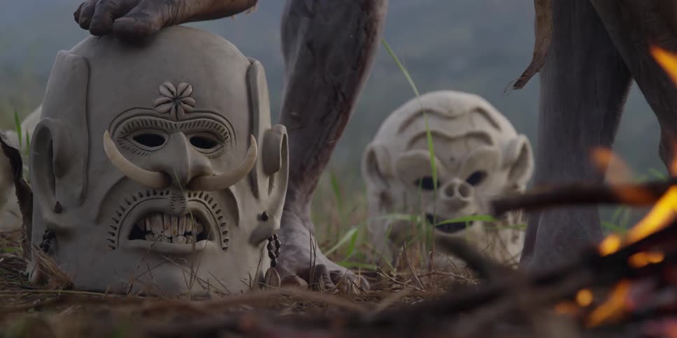 Men stand by a fire and very large clay masks in the forest