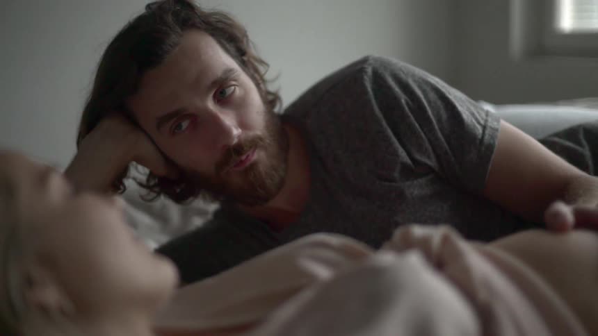 A man is lying next to a woman on a bed with his hand on her bare pregnant stomach