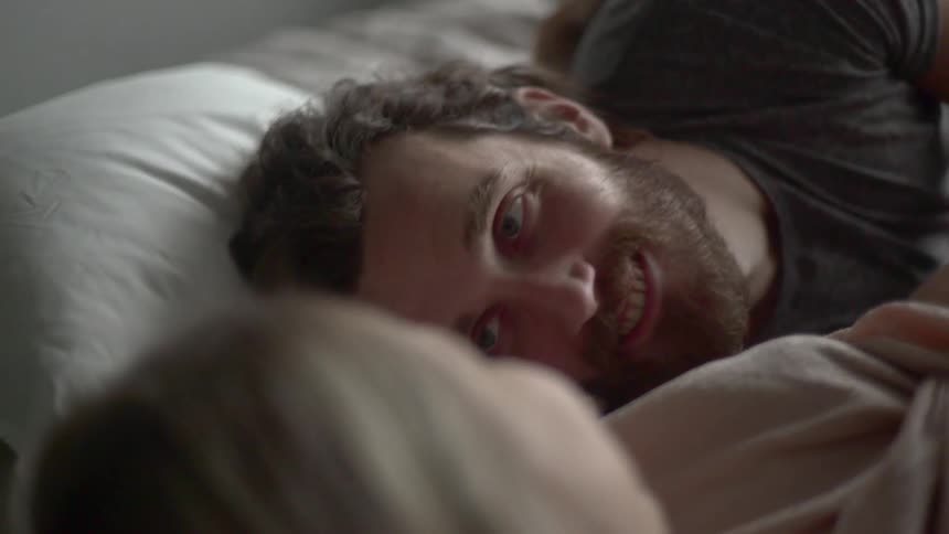 A bearded male puts his ear to womb of his pregnant partner before looking up to her at smiling as they lay on a bed