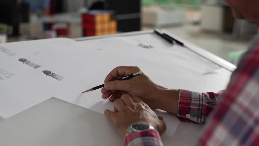 A man sits at a desk sketching on paper with a pencil