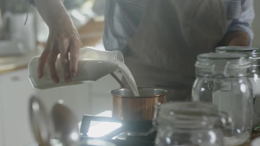 A cook pouring milk from a glass bottle into a sauce pan