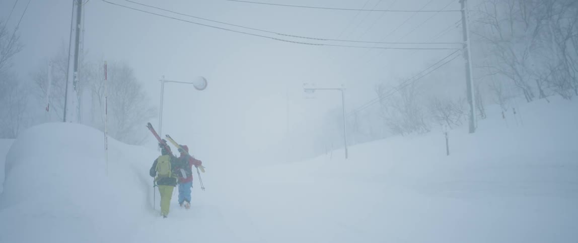 A couple of people walk with their skis resting on their shoulders along a path covered in heavy snowfall