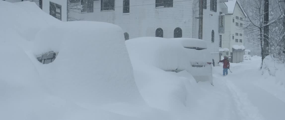 A person carrying skis and poles walks past building through snow